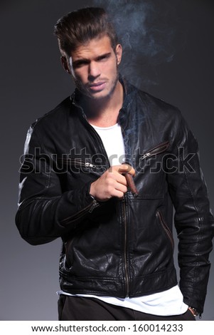 young man in leather jacket is blowing the smoke and looks at the camera