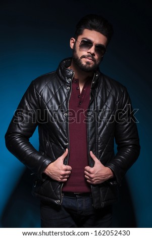 young man with long beard is holding his leather jacket and looks at the camera