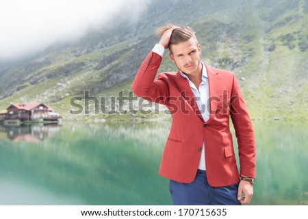 young man fixing his hair by passing his hand though it in front of a mountain lake with cabin