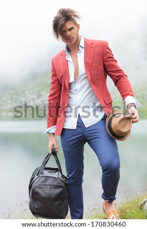 young man with blown hair is standing in front of a lake and looks at the camera while holding his bag and hat