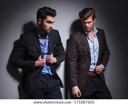 two hot male models posing against studio background, one looking away and one pulling his suit\'s collar