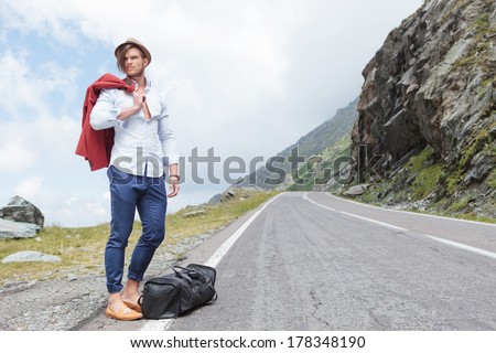 attractive young fashion man posing outdoor, on the side of the road, with his jacket over his shoulder and his bag at his feet, while looking away