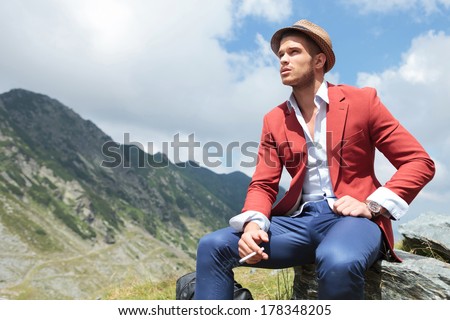 picture of a young fashion man sitting on a rock in the mountains, while looking away from the camera with a cigarette in his hand