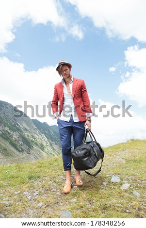 full length picture of a young fashion man walking in the mountains with a bag in his hand while looking into the camera