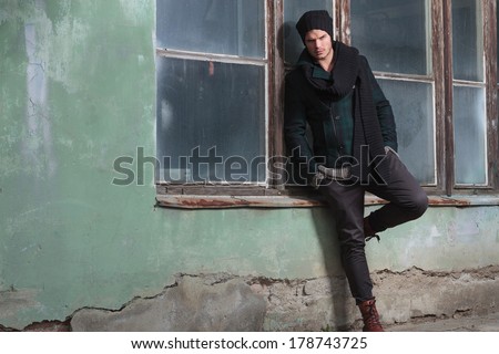 young fashion man leaning on a window ledge / sill while holding his hands in his pockets and looking into the camera