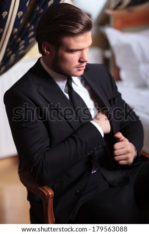elegant young business man sitting on a vintage chair and introducing his hand inside his suit jacket while looking away from the camera, in a retro hotel room