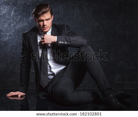 seated provocative elegant fashion man is fixing his black tie in studio