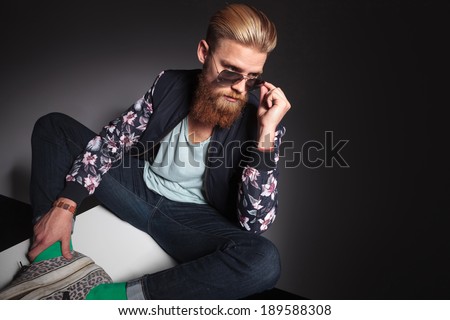 young bearded man looking over his sunglasses while sitting with his feet together. on a black studio background