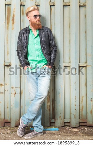 portrait of a young casual redhead bearded man standing outdoor with his crossed feet and his hands in his pockets while looking away from the camera