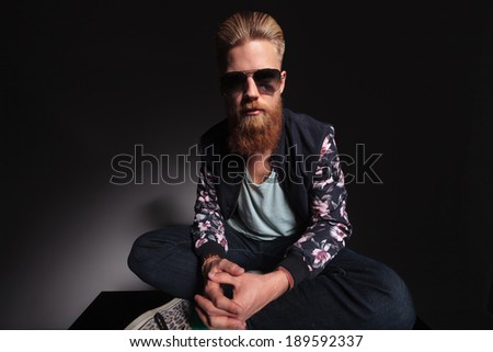 young bearded man looking into the camera while sitting with his feet crossed and his hands together. on a black studio background