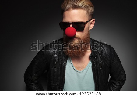 casual young man with a long beard, wearing a clown's red nose and looking away . on a dark studio background