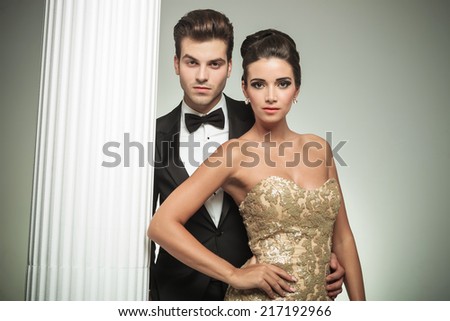 fashion elegant couple man in tuxedo and woman in evening gown standing embraced near column in studio