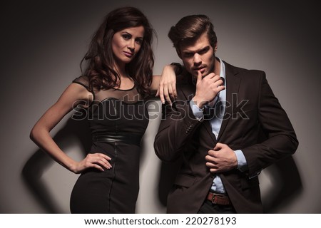 Young fashion couple looking into the camera while the man is pulling his jacket with one hand and touching his lip with his thumb. The woman is leaning on him. On dark grey background.