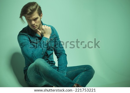Close up picture of a young fashion man looking down while sitting on the floor with his thumb at his chin.