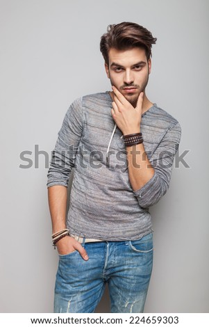 Portrait of a young fashion man holding his hand in pocket while fixing his beard, looking at the camera.