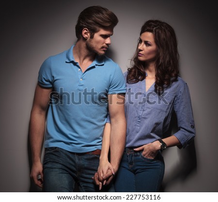 In love fashion couple looking at each other while holding hands, leaning on a grey wall.