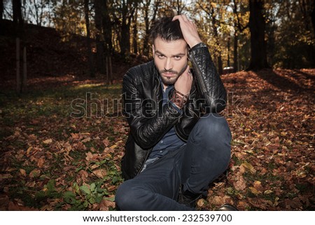 Fashion man sitting in the park, holding one hand in his hair and the other one to his chin.