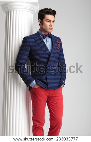 Picture of a handsome young fashion man posing near white column, looking away with both hands in his pocket.