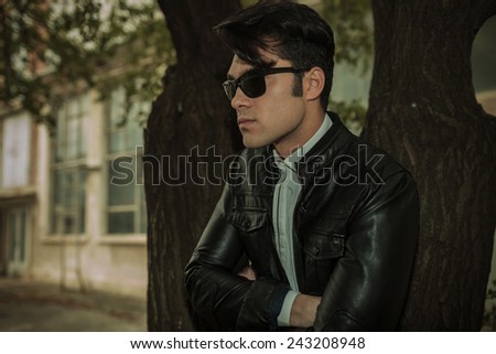 Close up picture of a handsome casual man leaning on a tree with his hands crossed, looking away from the camera.