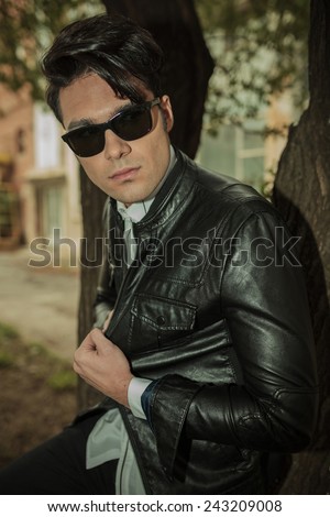 Handsome young fashion man pulling his leather jacket while leaning on a tree in the park, looking away.