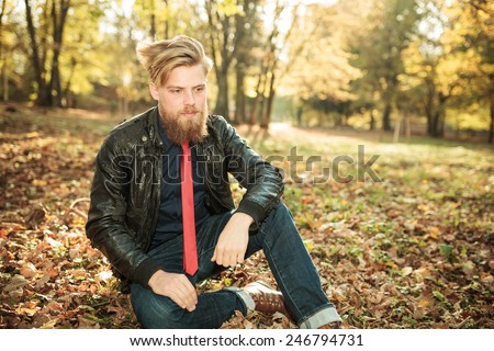 Young casual man looking down while sitting in the park, resting one hand on his knee.