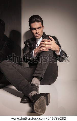 Young fashion man leaning on a wall while sitting on the floor, holding his knee up with both hands, looking away from the camera.