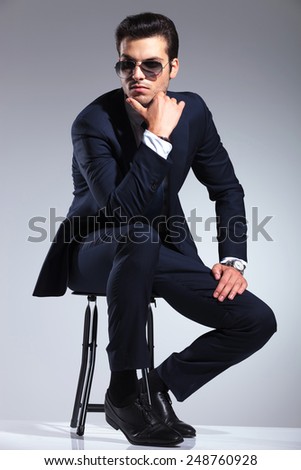 Full body picture of a young elegant business man sitting on a stool holding his hand to his chin, thinking.