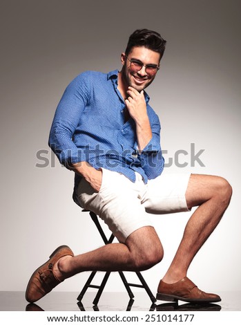 Side view of a happy young man sitting on a chair, holding one hand in his pocket while petting his beard.