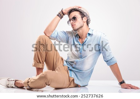 Fashion man holding his hat with his rigth hand while sitting on the floor, looking away.