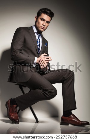 Side view picture of a young elegant business man sitting with one leg in front of the other, looking at the camera.