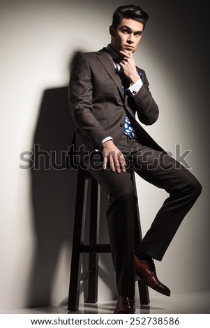 Elegant business man sitting on a stool against studio background, thinking while holding his hand to his chin.