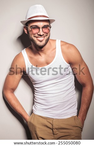 Smiling young fashion man leaning on a white wall holding both hands in his pocket.