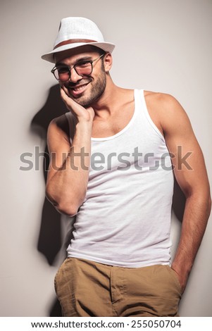 Happy young casual man leaning on a white wall with one hand in his pocket while holding the other hand on his cheek.