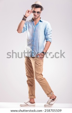 Full length picture of a young casual fashion man taking off his sunglasses while holding one hand in his pocket.
