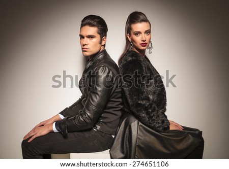 Fashion man and woman sitting back to back while holding their hands on the legs.