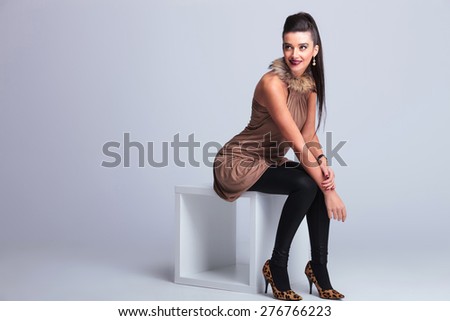 Full body picture of a young elegant fashion woman sitting on a chair while smiling and looking up.