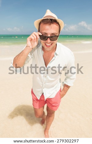 Full body picture of a handsome young man smiling to the camera while taking off his sunglasses.