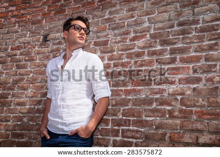Young casual man holding his hands in pockets while leaning on a brick wall, lloking away from the camera.
