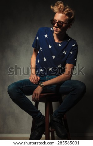 side view of a fashion man with messy hair and sunglasses sitting on a stool in studio