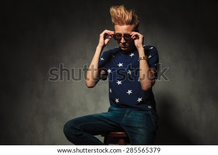 fashion man is taking off his sunglasses while sitting on a chair in studio