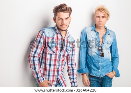 two young fashion men standing against studio wall and looking at the camera