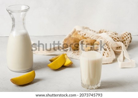 In a bottle and in a glass, the drink is vegetable, vegetable potato is not milk. Nearby there are potatoes cut into wedges, on a white background, and a knitted string bag with whole potatoes