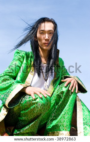 young asian man in green Mongolian suit with his hands on knees looks into camera