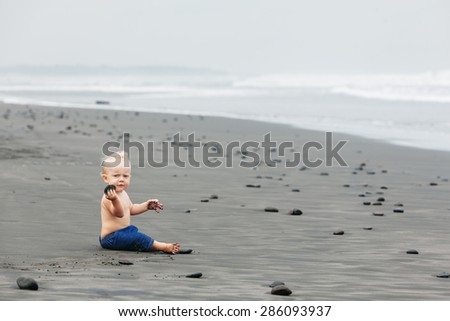 Portrait of smiling child sitting and playing alone with fun on balinese black sand sea beach before swimming Family lifestyle, water leisure during summer vacation with baby on tropical Bali island