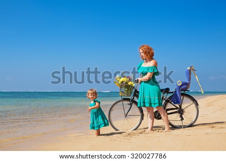 Happy family - baby girl with young mother after bicycle exercises walk with bike and have a fun on sea sand tropical beach. Active parents and people outdoor activity on summer vacation with children