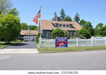 American Flag Sold (Another success let us help you buy sell your next home) real estate sign on front yard lawn of suburban home with white picket fence clear blue sky USA residential neighborhood