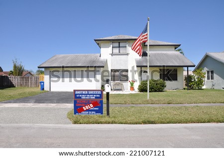 American Flag pole Real estate (another success let us help you buy sell your next home) sign on front yard lawn of suburban home residential neighborhood USA clear blue sky