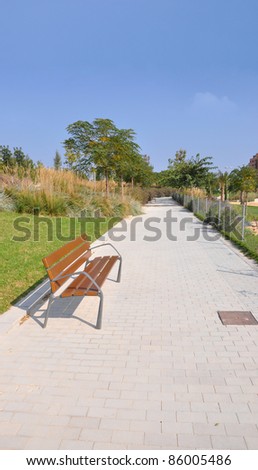 Empty Wood Park Bench on Brick Path lined with Grass Plants and  Trees on Sunny Blue Sky Day