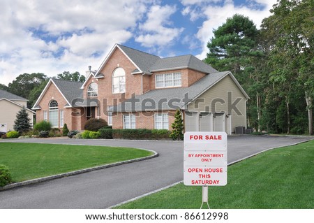 Realtor For Sale Sign on Front Yard Lawn of Large Three Car Garage Suburban Brick McMansion Home in Residential Neighborhood on Cloudy Blue Sky Day