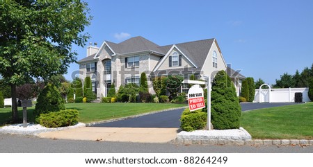 Realtor For Sale Sign on front yard of beautiful landscaped large brick suburban home on sunny clear blue sky day
